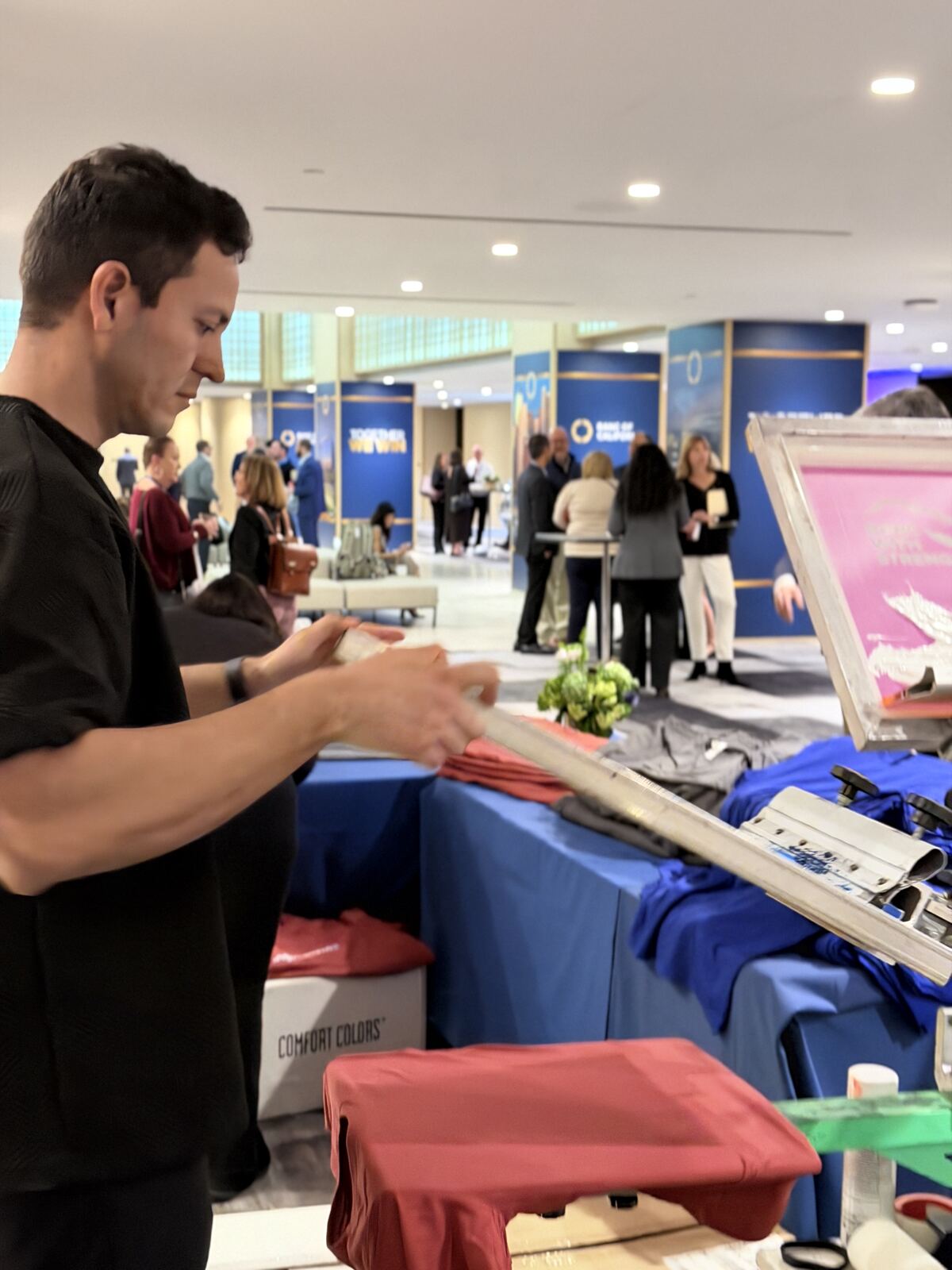 Staff member operating a trade show booth displaying blue and red printed tshirts with samples