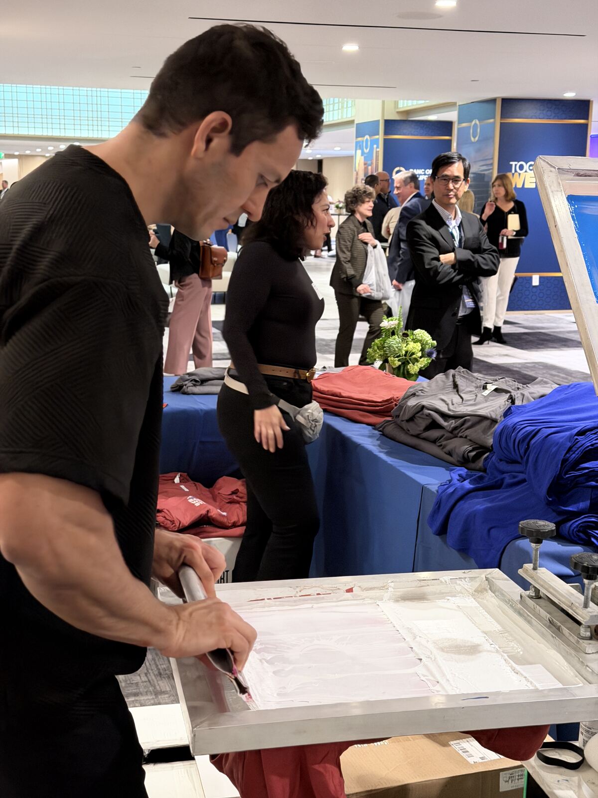 Young man demonstrating screen-printing technique on apparel at a trade show booth with visitors observing